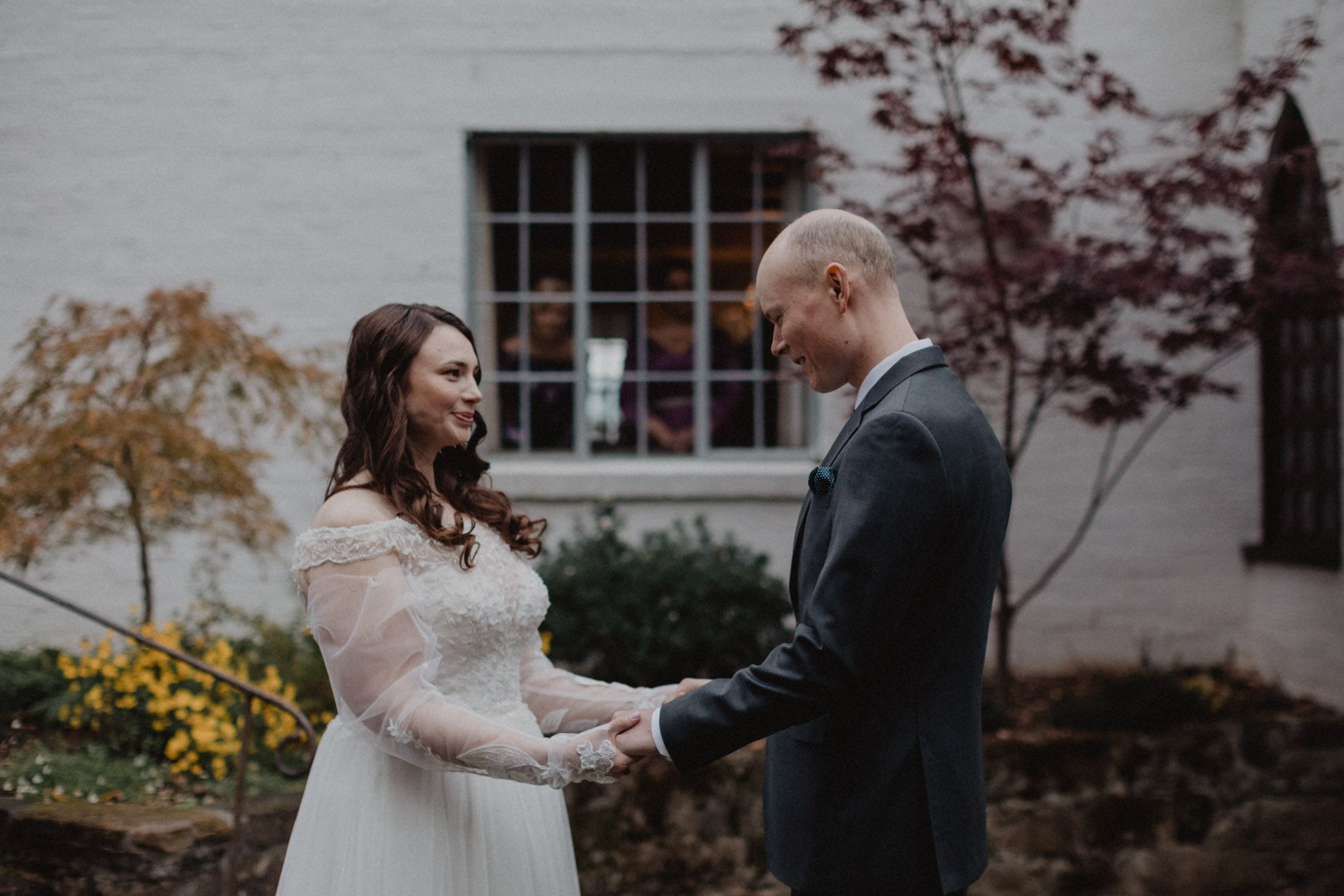 Bride and groom embrace during their first look in front of a white building with autumn foliage.