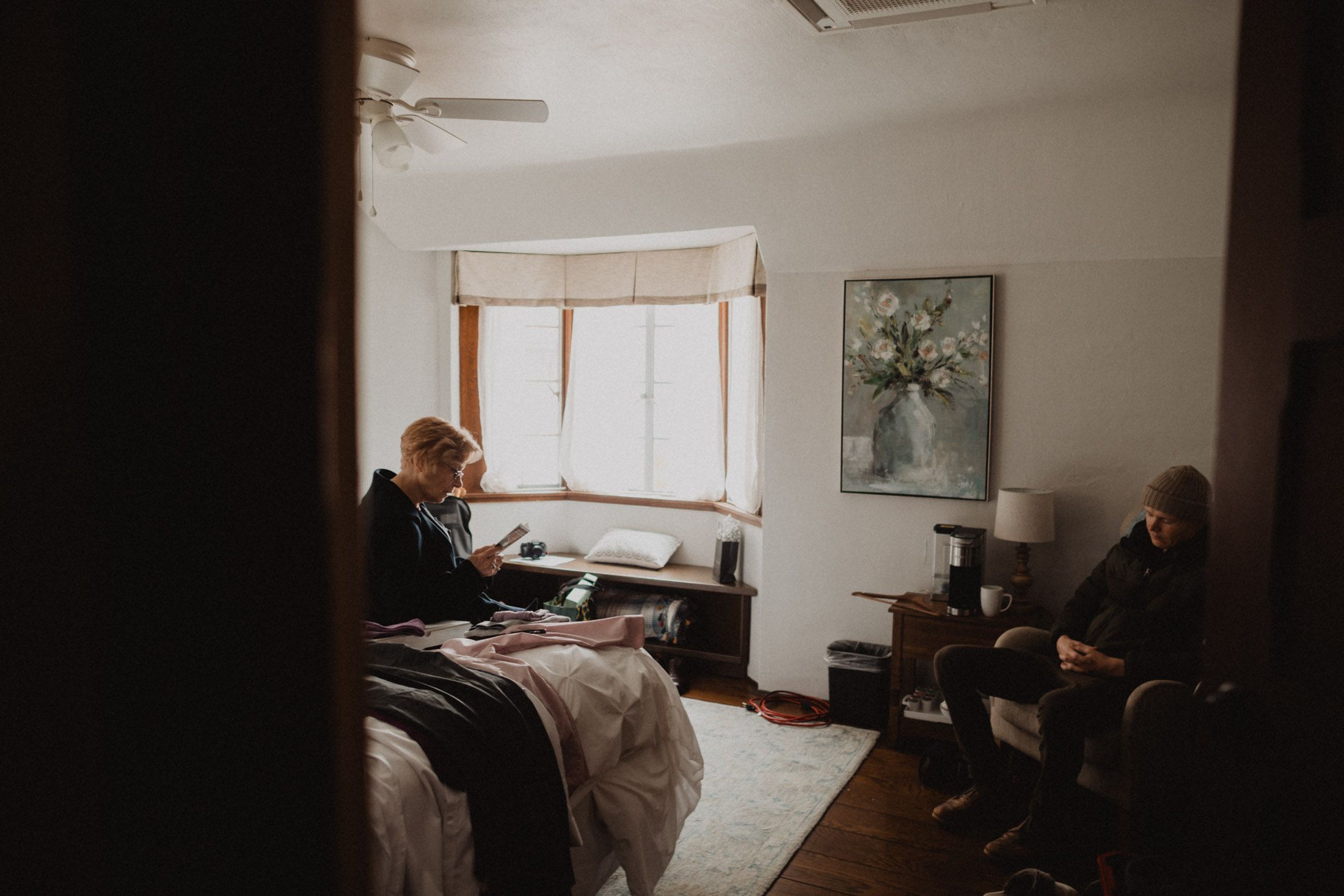 A moody interior bedroom scene with two people sitting in natural window light and vintage decor.