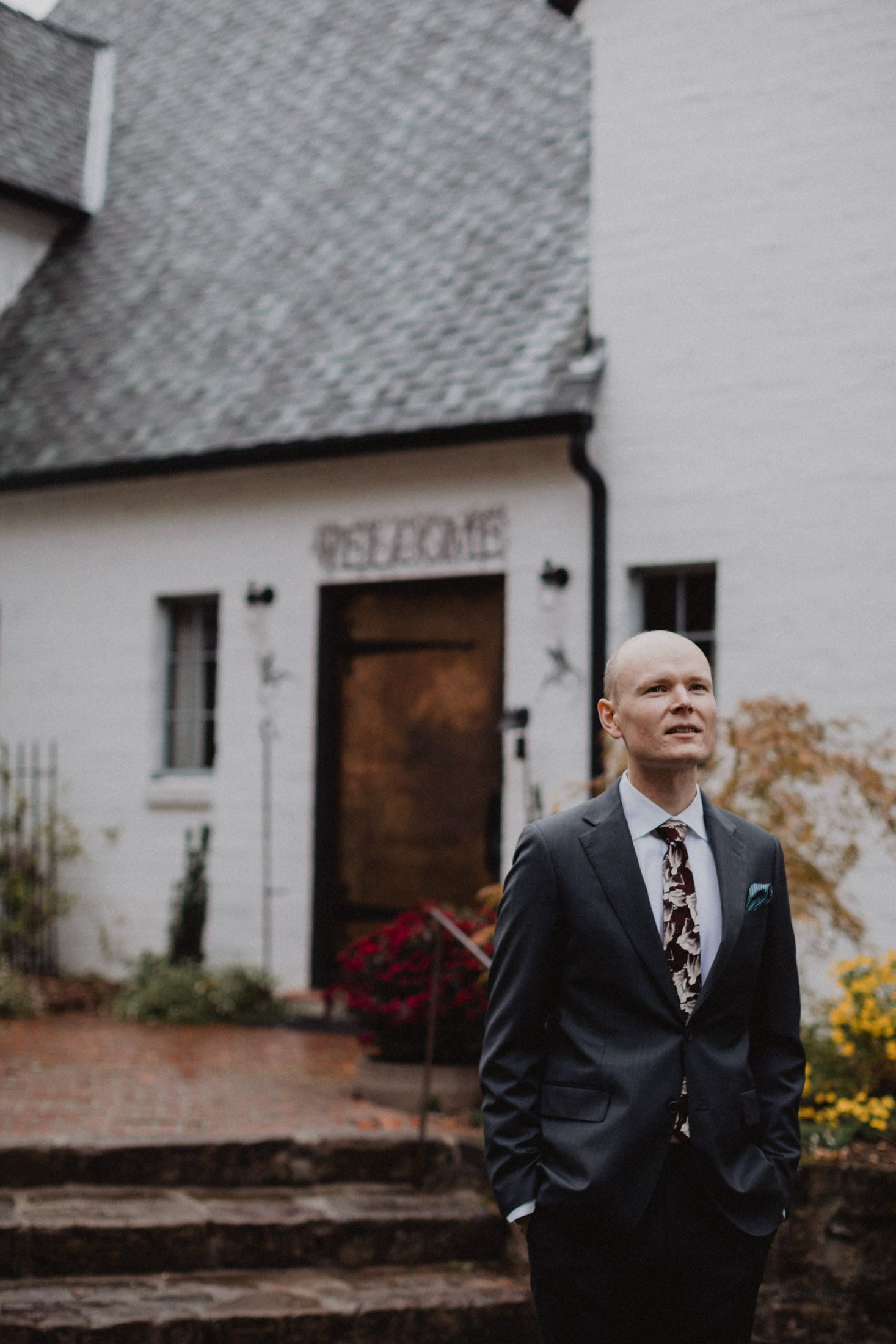 A person in a dark suit and patterned tie stands outside a white building with stone steps and a wooden door.