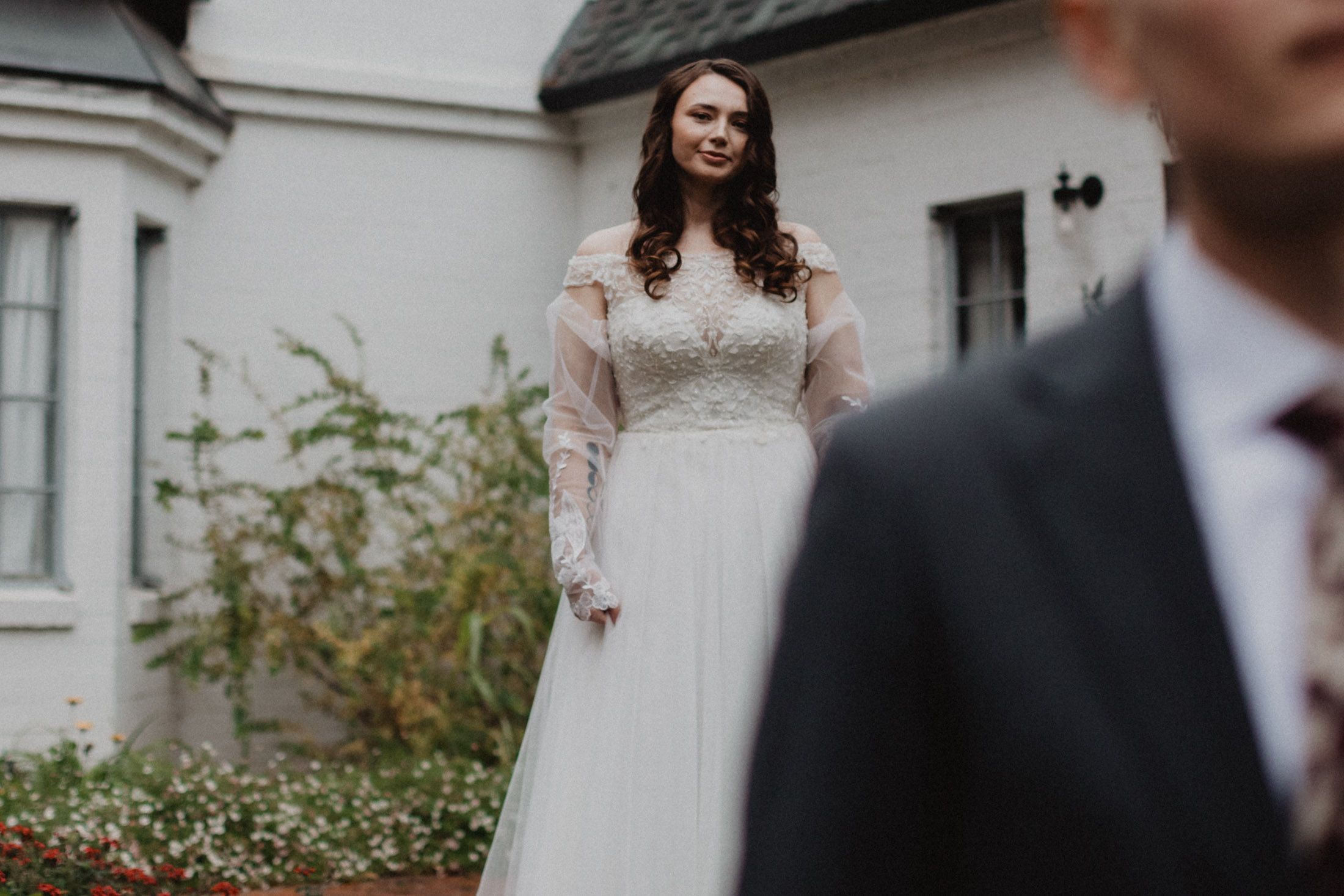 A bride in a long-sleeved white lace wedding dress stands outside a white building while someone in a suit is visible in foreground.