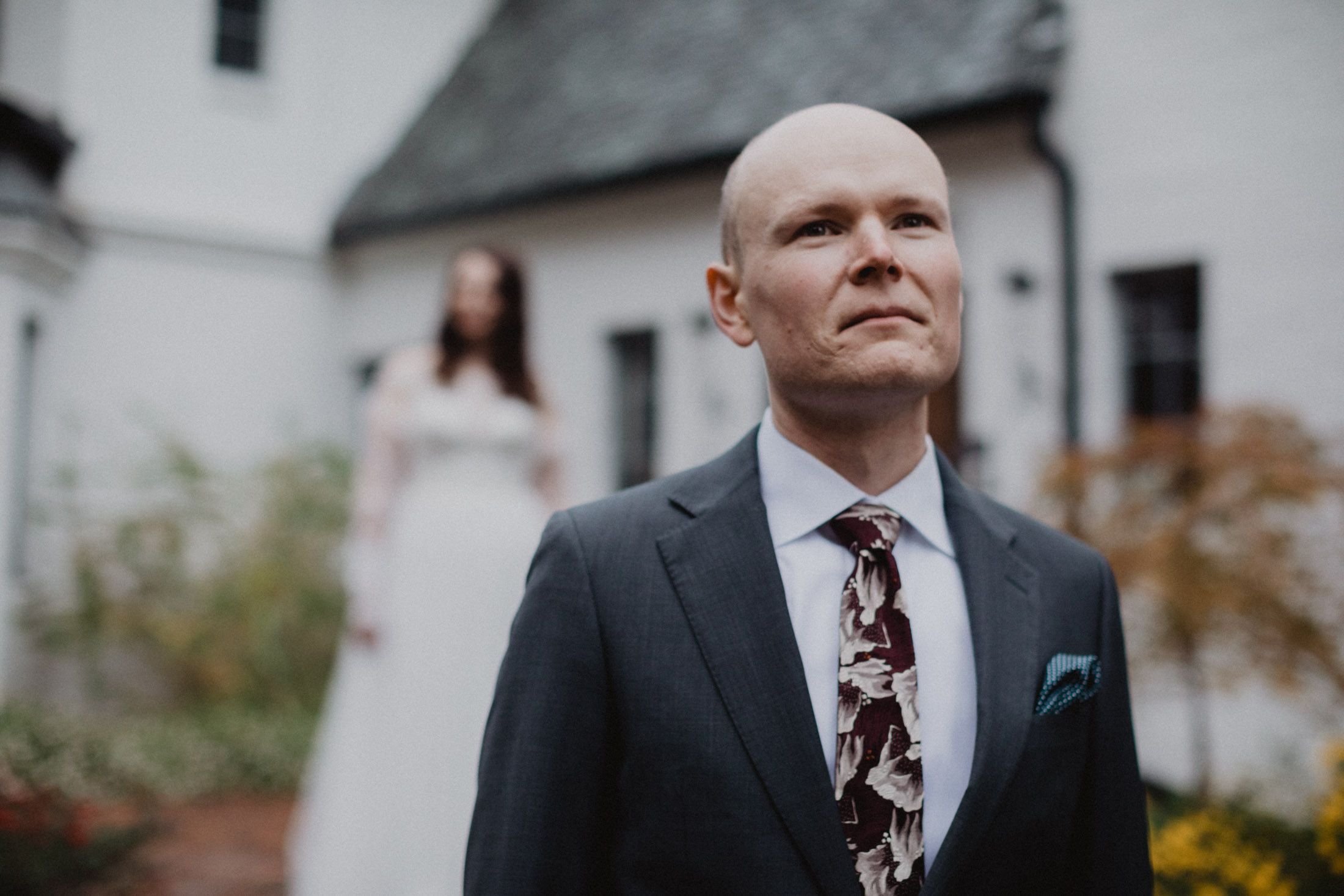 A group of formally dressed people gather outdoors for a wedding celebration at a white building.