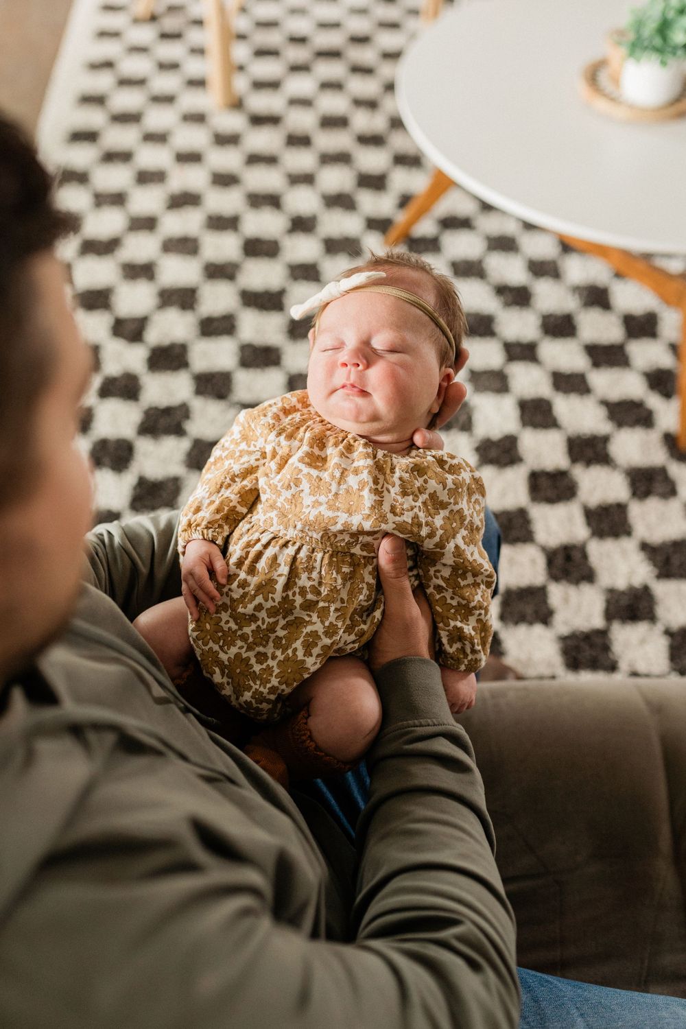 A baby in a floral outfit lies on a couch while being held by someone wearing a green jacket.