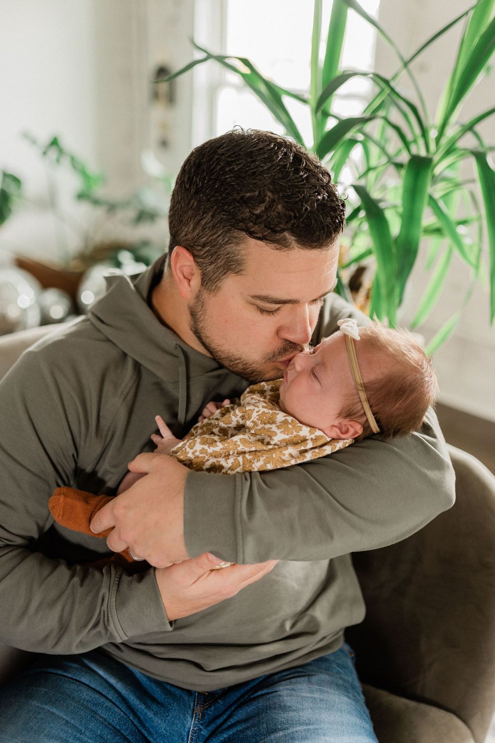 A tender moment between parent and sleeping baby on a couch, with houseplants visible in the background.