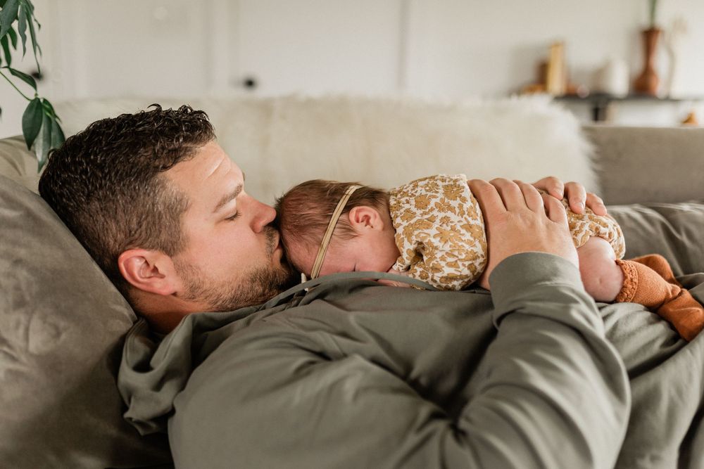 A parent and baby share a tender moment while resting together on a gray couch.