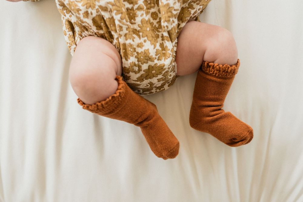 Baby wearing brown ruffle-top socks and floral dress lying on white bedding.
