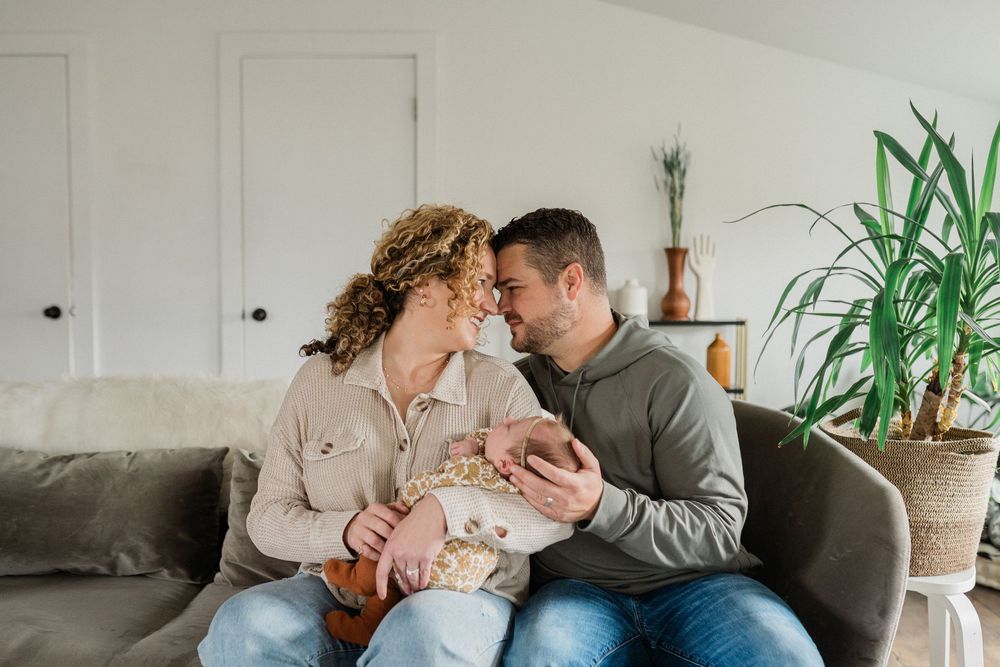 A couple sits together on a gray couch holding a newborn baby while indoor plants decorate their bright living room.