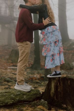 Couple kissing in misty forest, girl standing on tree stump in floral dress and sneakers, boy in burgundy sweater.