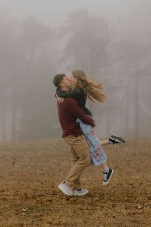 A couple sharing a romantic kiss in a foggy autumn park, man lifting woman as her hair flows freely.