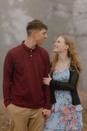 Young couple holding hands and gazing at each other, woman in floral dress smiling, man in maroon shirt, outdoors.