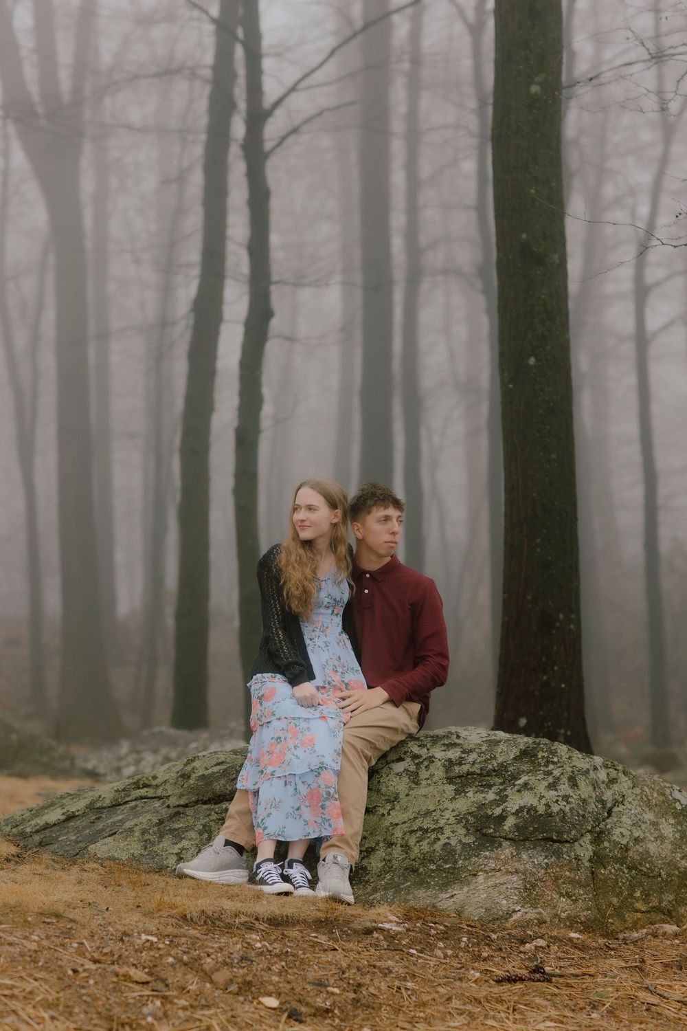 Couple sitting on a mossy rock in a misty forest, woman in floral dress and man in burgundy sweater.