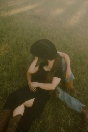 Two people sharing a romantic embrace in a sunlit green field, one wearing a black cowboy hat and boots.