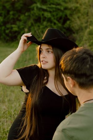 Young woman in black cowboy hat and shirt sitting outdoors in golden hour light with a companion nearby.