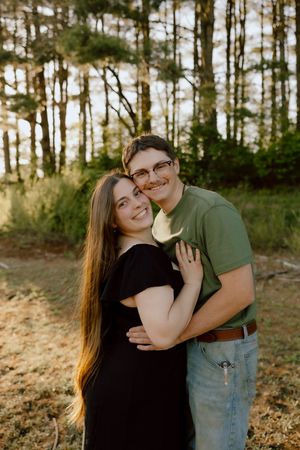 A smiling couple embraces outdoors in a sunlit forest, the man in a green shirt holding a white flower.