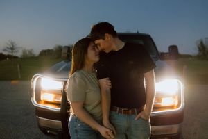 Romantic couple standing in front of illuminated truck headlights at dusk, sharing an intimate moment outdoors.