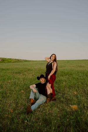 A couple poses in a green field at sunset, man in cowboy hat sitting while woman in red skirt stands behind him.