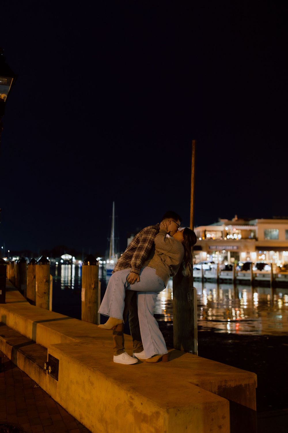 Couple sharing a kiss on a waterfront dock at night with marina lights reflecting on the water.