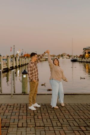 Joyful couple dancing on a brick waterfront promenade at sunset with a peaceful harbor behind them.