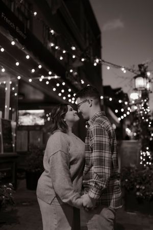 Couple holding hands and sharing a kiss under string lights on a charming city street at night.