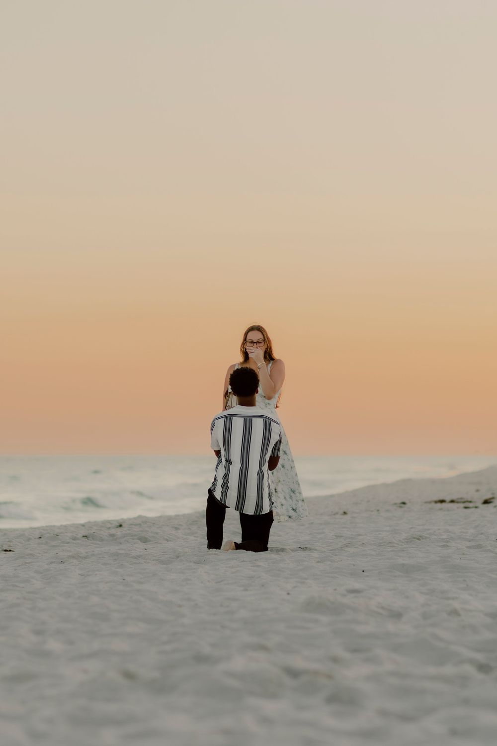 Man kneeling on sandy beach proposing to woman at sunset with ocean waves in background.