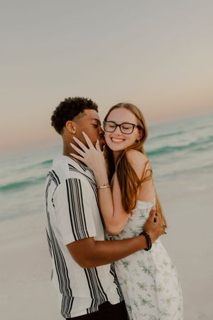A young couple embraces and shares a kiss on a beach at sunset, with turquoise ocean waves in the background.