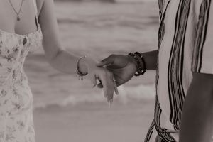 Black and white photo of a couple holding hands on a beach, wearing bracelets, with ocean waves in the background.
