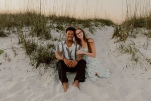 Couple sitting together on white sand beach dunes at sunset, smiling and embraced in a romantic outdoor photo session.