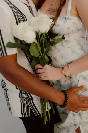 Couple embracing while holding a bouquet of white roses, wearing a striped shirt and floral dress.