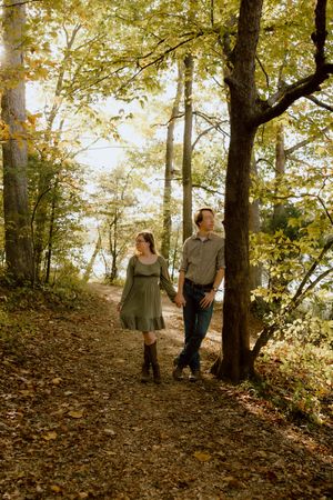 Couple holding hands while walking along a scenic autumn forest trail surrounded by golden foliage.