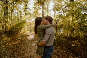 Couple sharing a romantic kiss on a sunlit forest path surrounded by golden autumn leaves.