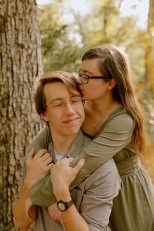 Young woman kissing a smiling man on the cheek while hugging him from behind near a tree.