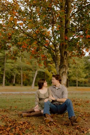 Couple sitting together under a colorful autumn maple tree surrounded by fallen leaves in a park.