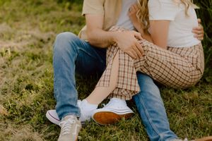Couple sitting together on green grass, wearing jeans and casual outfits, sharing a tender embrace outdoors.