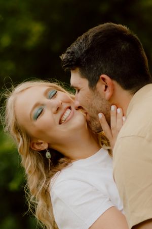 A smiling blonde woman with teal eye makeup receives a kiss on the cheek from a dark-haired man outdoors.