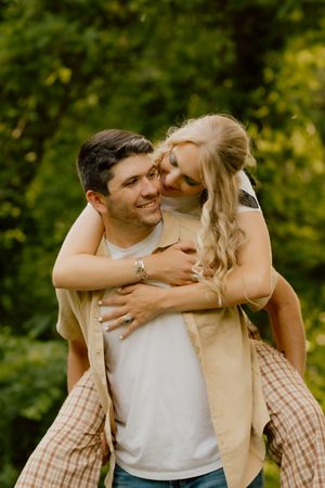 A smiling man gives a laughing blonde woman a piggyback ride outdoors surrounded by lush green trees.