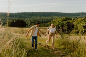 A couple holds hands running through a sunny green meadow with rolling hills and vineyards in the background.