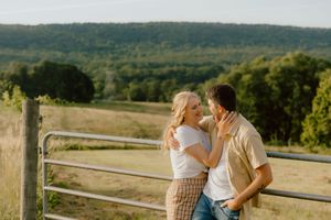 Couple embracing and smiling at each other by a farm fence with rolling green hills in the background.