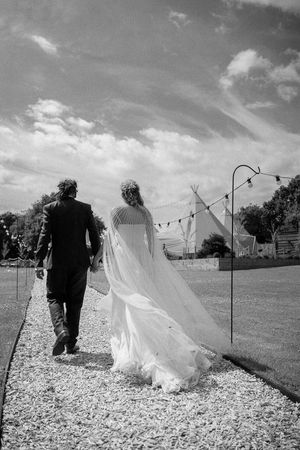 A couple walks along a gravel path at an outdoor wedding venue with string lights and cloudy skies overhead at the tipis at riley green.