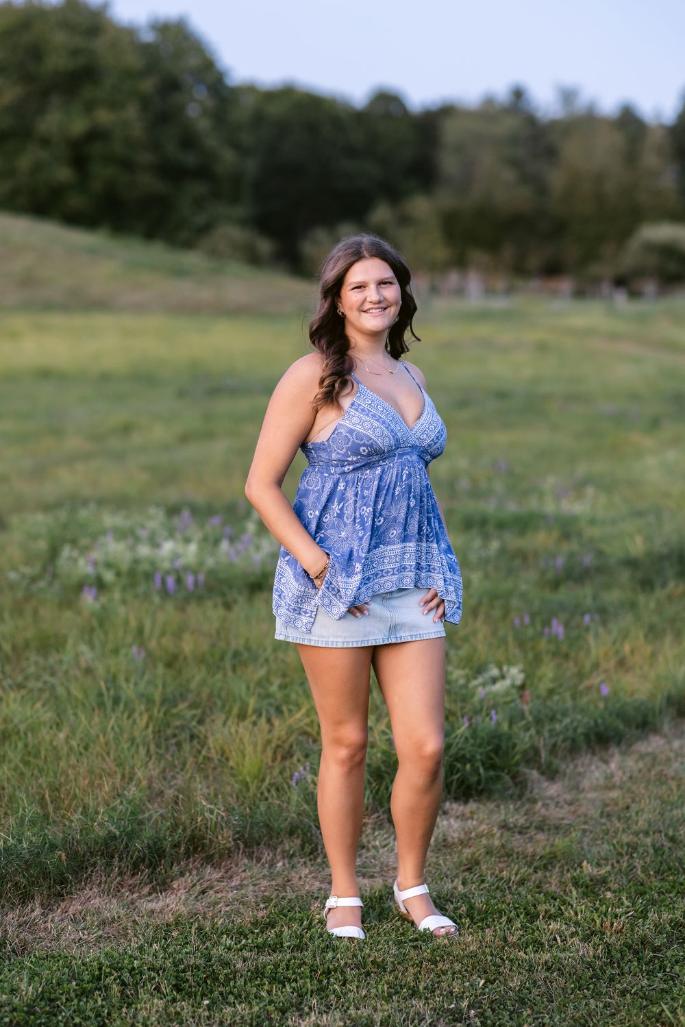 A senior girl wearing a blue handkerchief shirt and jean shorts stands in a field at Stevens Coolidge Estate in North Andover, MA.