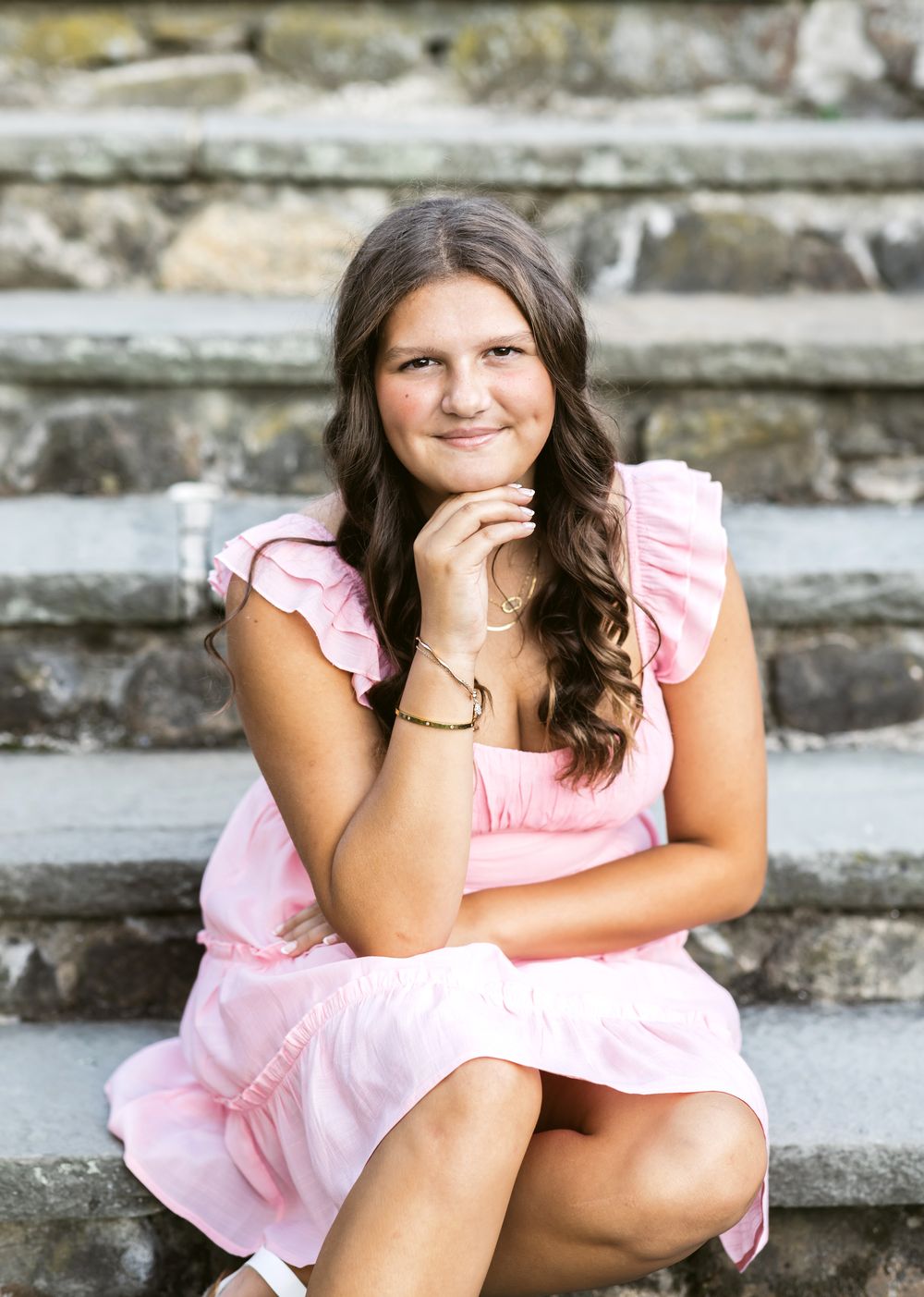 A senior girl wearing a pink dress sits on stone steps for senior photos at Stevens Coolidge Estate in North Andover, MA.