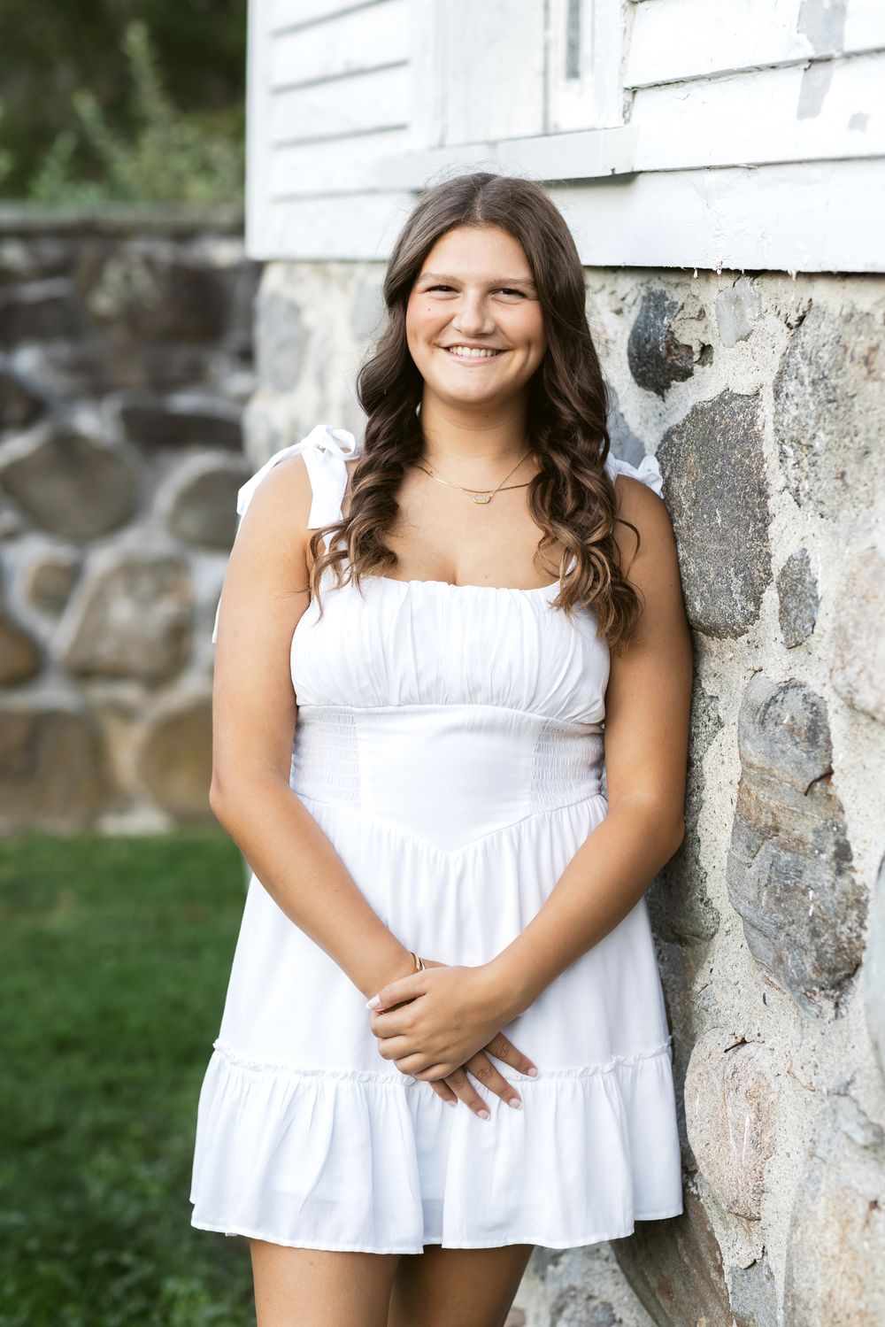 A young lady wears a white dress leaning a white farmhouse with a stone foundation at Stevens Coolidge Estate in North Andover, MA.