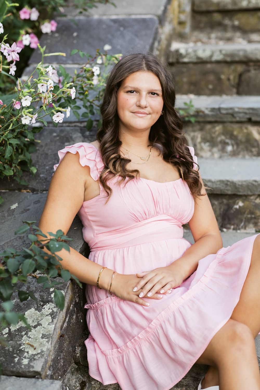 A girl with long brown hair wearing a pink dress sits on stone steps at Steve Coolidge Estate in North Andover, MA for senior photos.