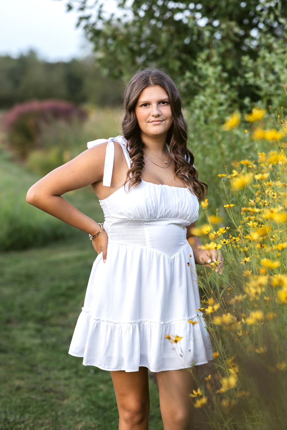 A senior girl with long brown hair wearing a white dress poses next to yellow flowers in a field at Stevens Coolidge Estate in North Andover