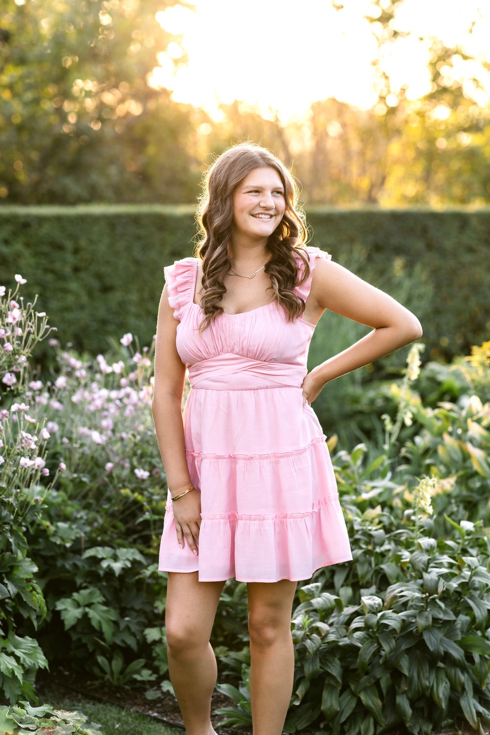 A senior girl wearing a pink dress poses with her hand on her hip in a garden at Stevens Coolidge Estate in North Andover for senior photos.