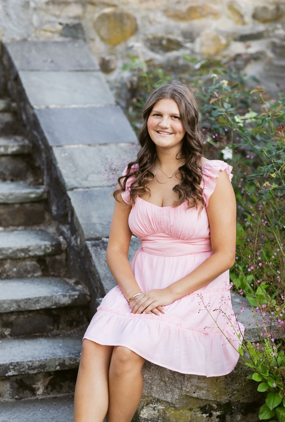 A senior girl with long brown hair wears a pink dress and sits on stone steps in a garden at Stevens Coolidge Estate in North Andover, MA.