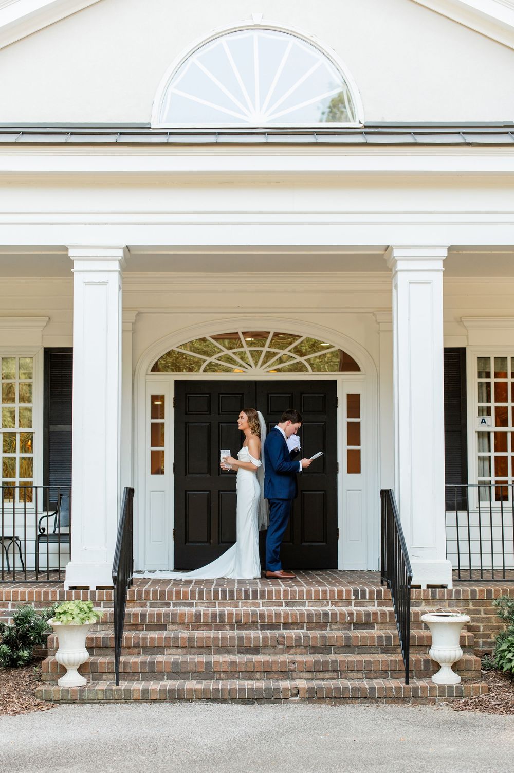 A wedding couple stands on the steps of a classic white colonial building with black doors.