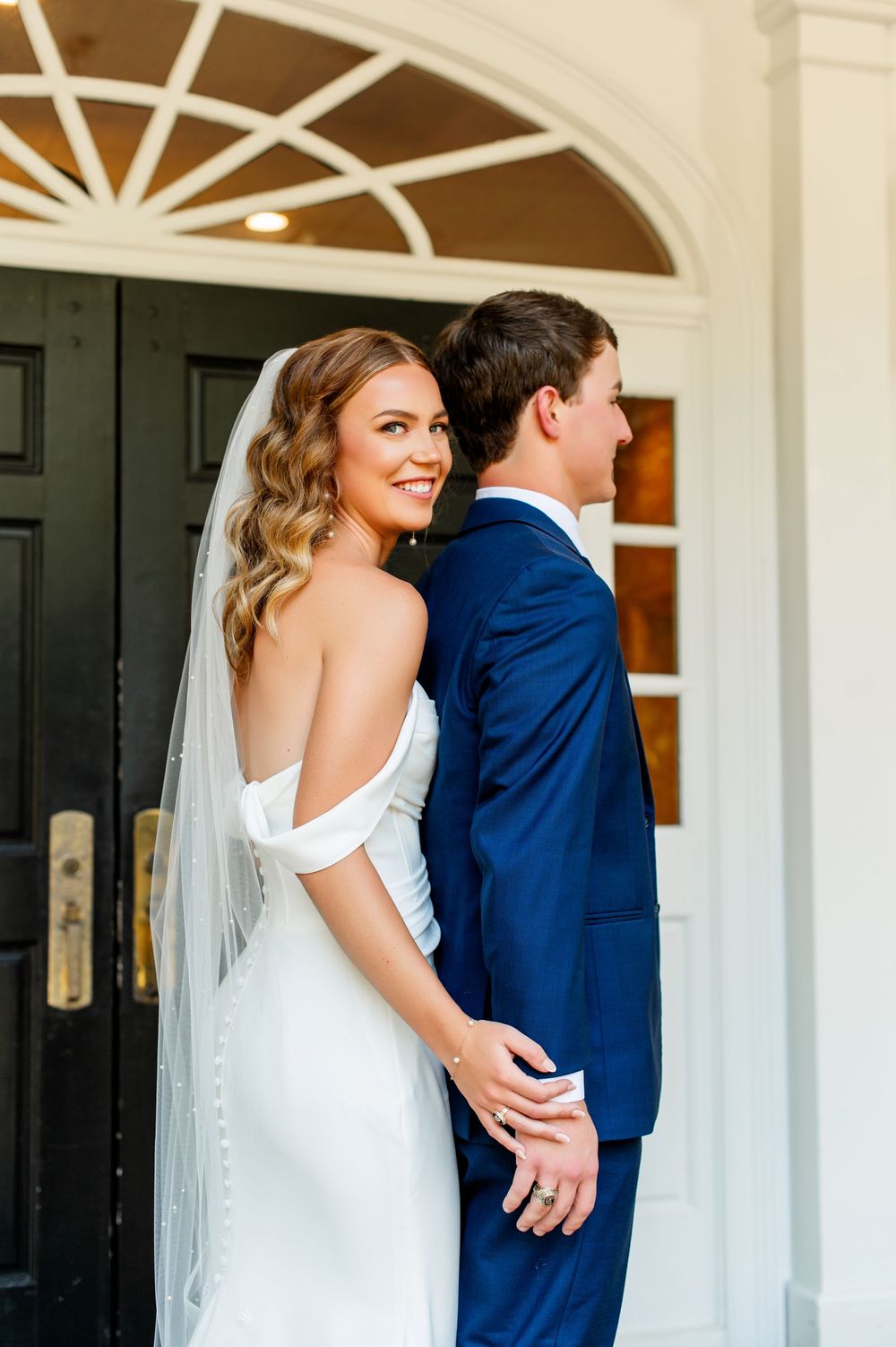 A couple in wedding attire poses together on a white porch with elegant architectural details.
