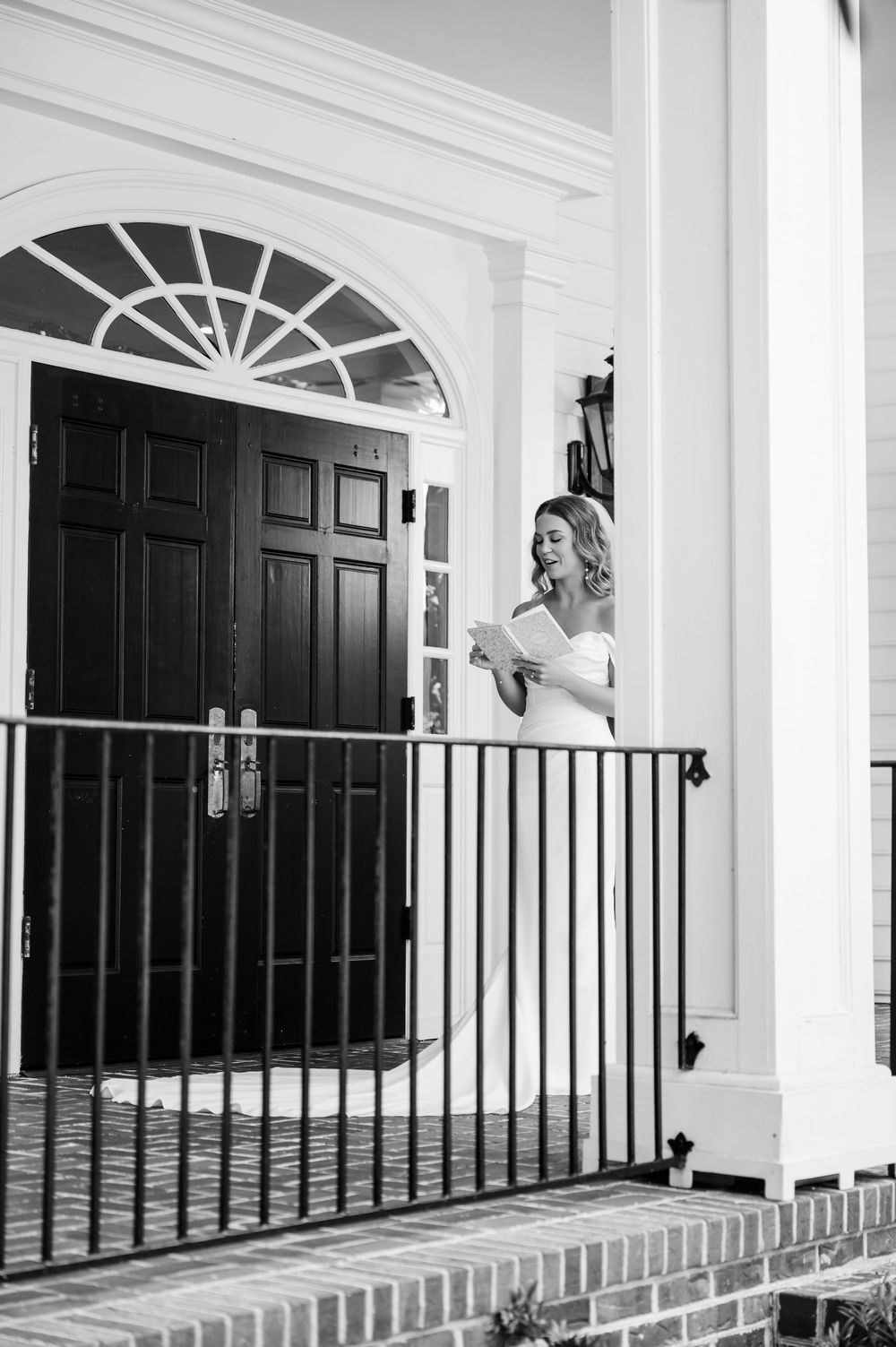 A black and white photo of a bride smiling on a white colonial porch with iron railings.