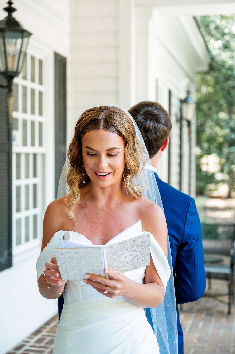 A bride in a white dress reads from a book while standing on a classic southern porch.