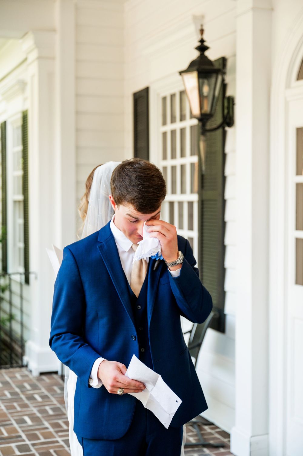 A groom in a navy suit wipes away tears while reading a letter on his wedding day.