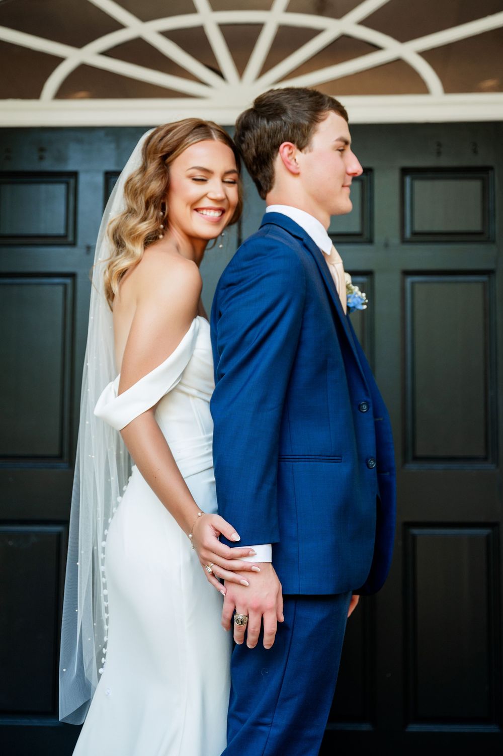 Wedding couple in white dress and navy suit pose against a dark wall with decorative trim.